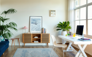 A bright home office with a large window, featuring a white desk with a computer and books, a wooden cabinet, and indoor plants. there's also a framed picture on the wall and a blue armchair.